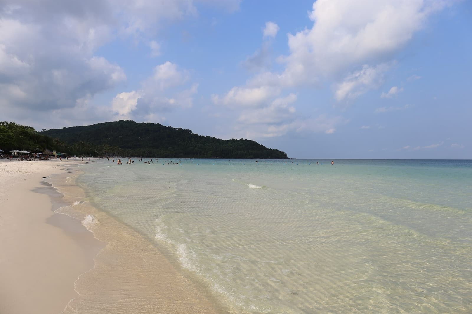 Shallow water clear enough to show the rippled sand beneath it at Bai Sao beach on Phu Quoc island, with swimmers offshore and a forested headland closing the bay