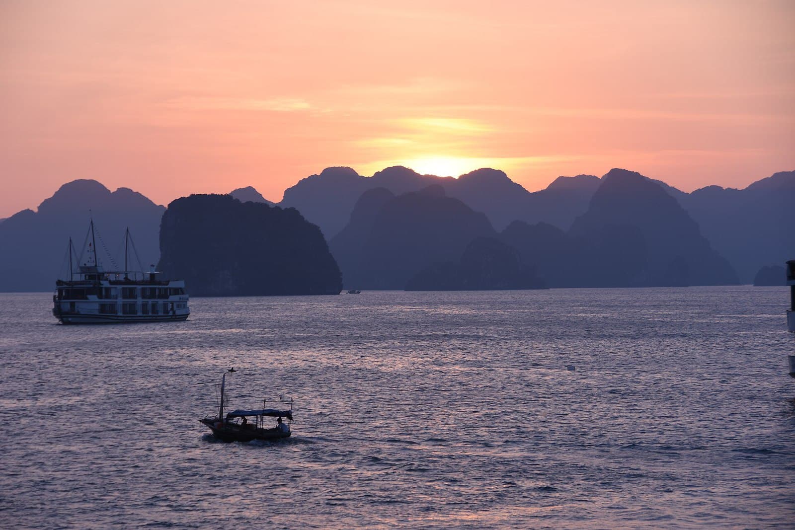 Sunset over Ha Long Bay — the sun dropping behind layered limestone karst islands, a white overnight cruise boat riding at anchor to the left and a small wooden service boat crossing the pink water in the foreground
