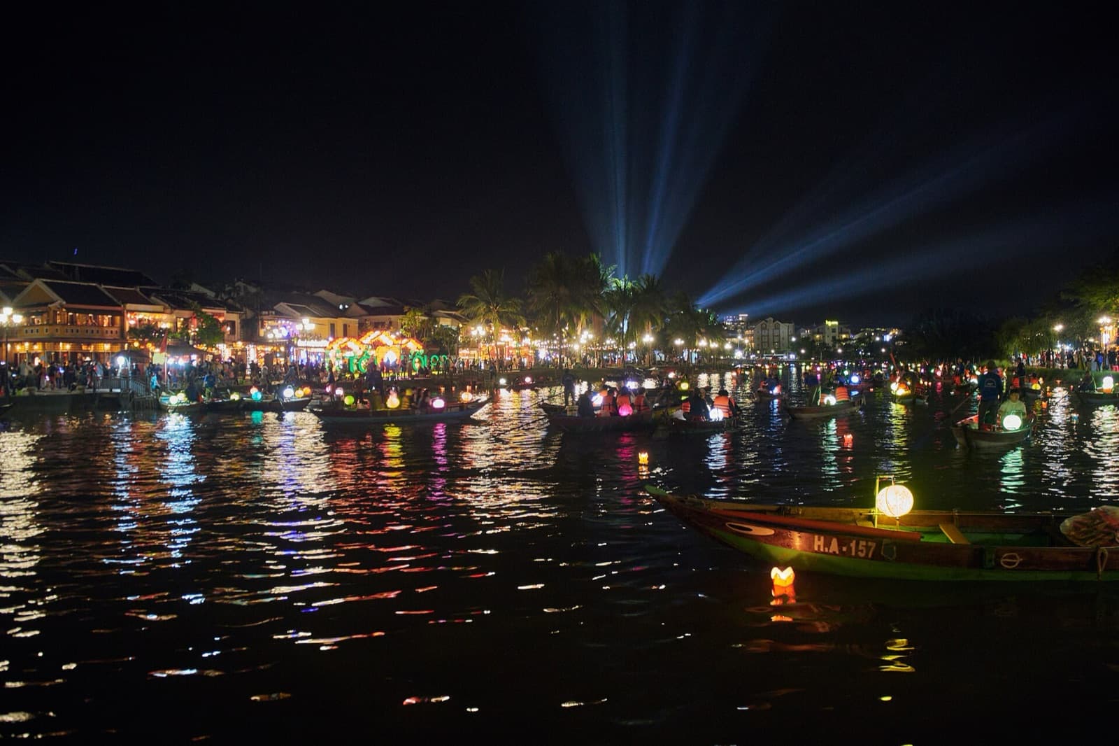 Rowing boats carrying lit paper lanterns on the Thu Bon River during a lantern night in Hoi An, with floating lanterns drifting on the water and the old-town riverfront lit behind