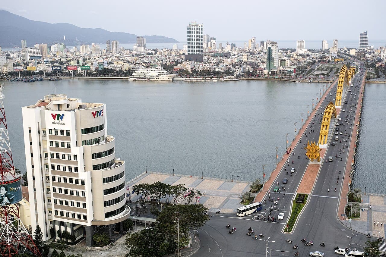 Da Nang seen across the Han River — motorbikes and cars streaming over the golden-pillared bridge toward a skyline of high-rises, with the coastal hills and the sea behind, at the southern end of the Hanoi to Da Nang run