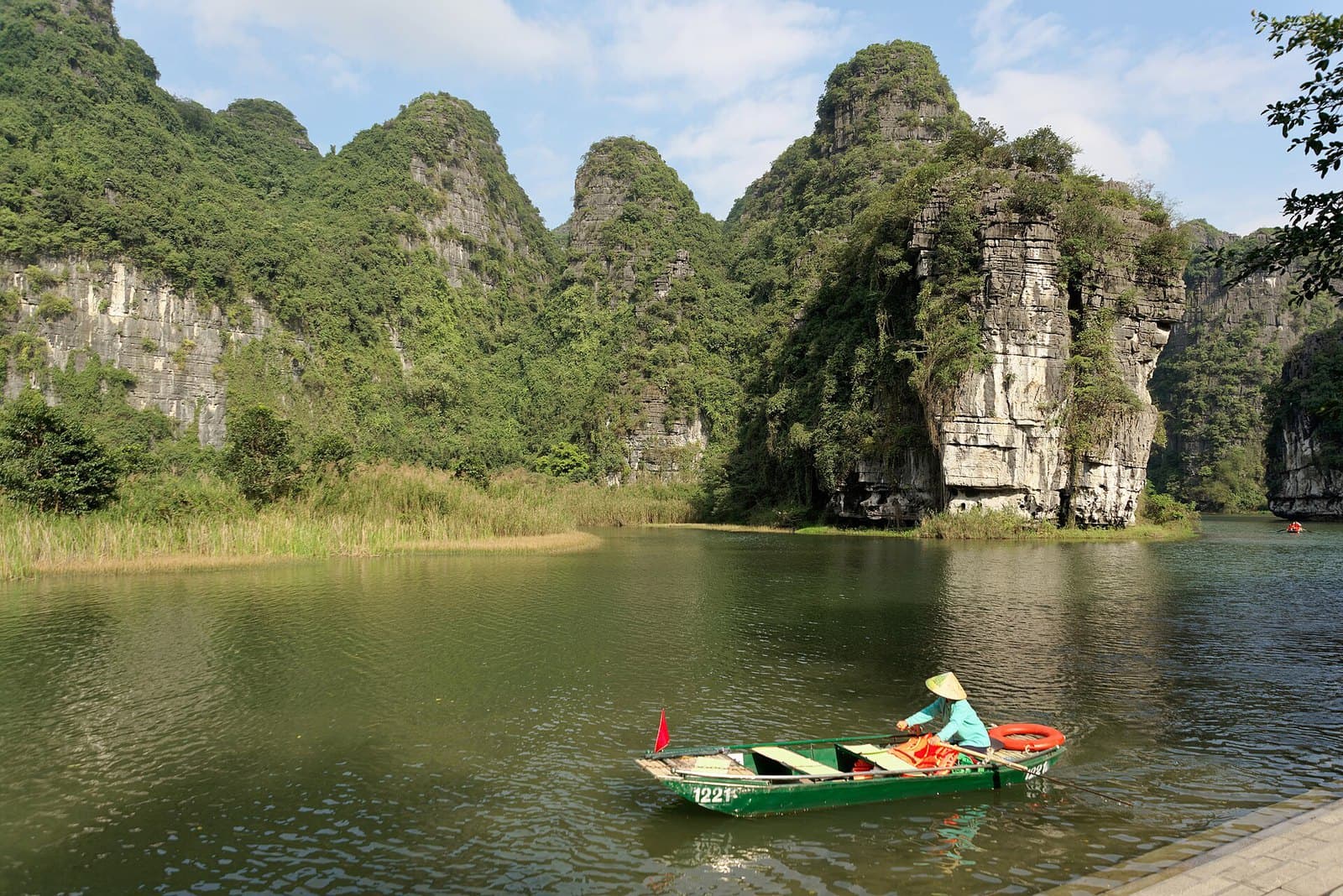 Forested limestone karst towers rising straight out of the green Trang An river in Ninh Binh, with a boatwoman in a conical hat rowing an empty green sampan across the open water